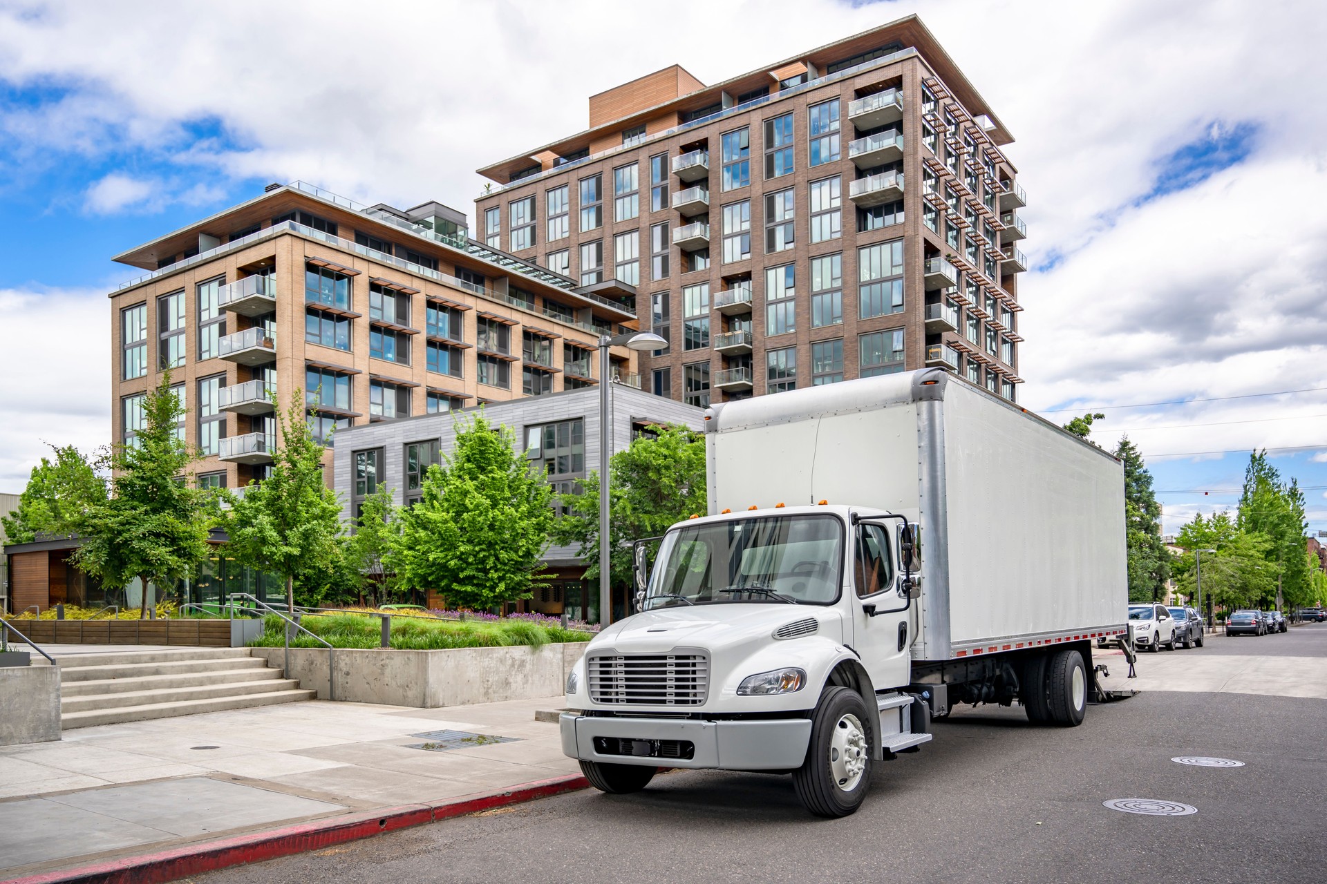 Popular local delivery and moving day cab middle duty semi truck with box trailer standing on the urban city street with multilevel apartment building unloading delivered goods